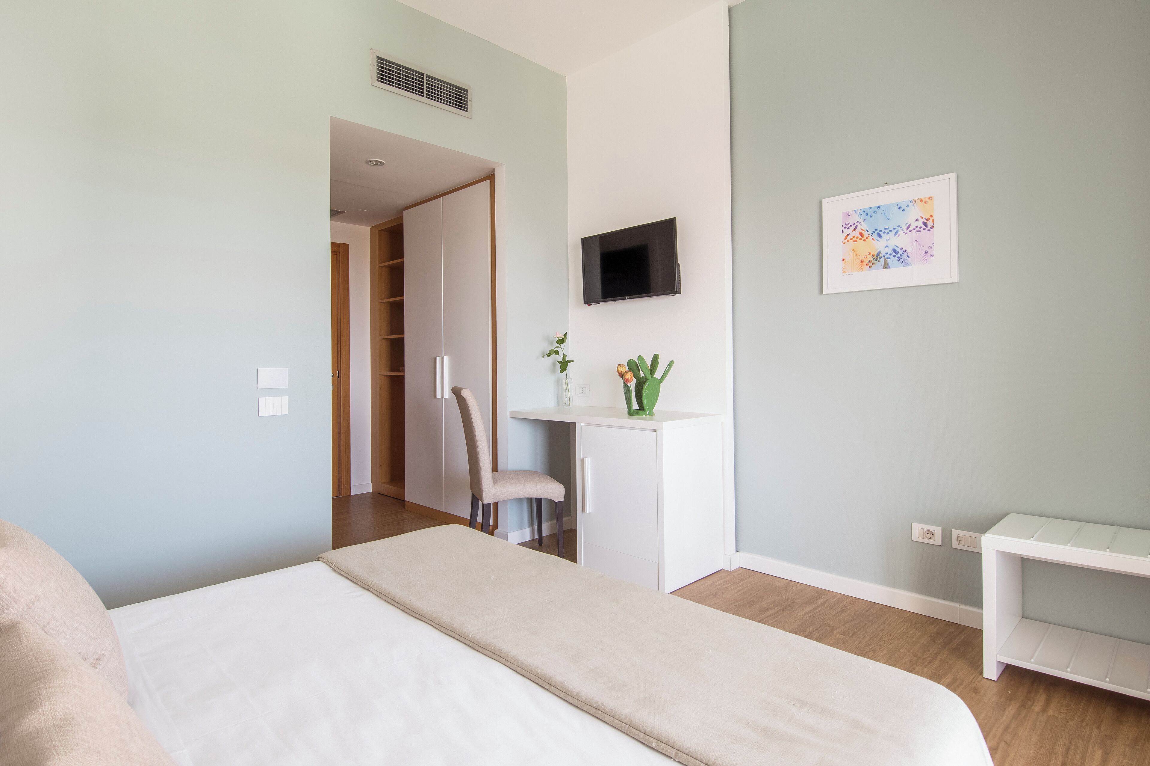 A modern bedroom with a bed, a desk, a chair, a TV, and a small plant.