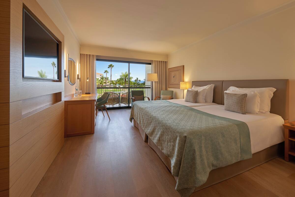 A hotel room with a large bed, a desk, a chair, a balcony with a view of palm trees, and a window.