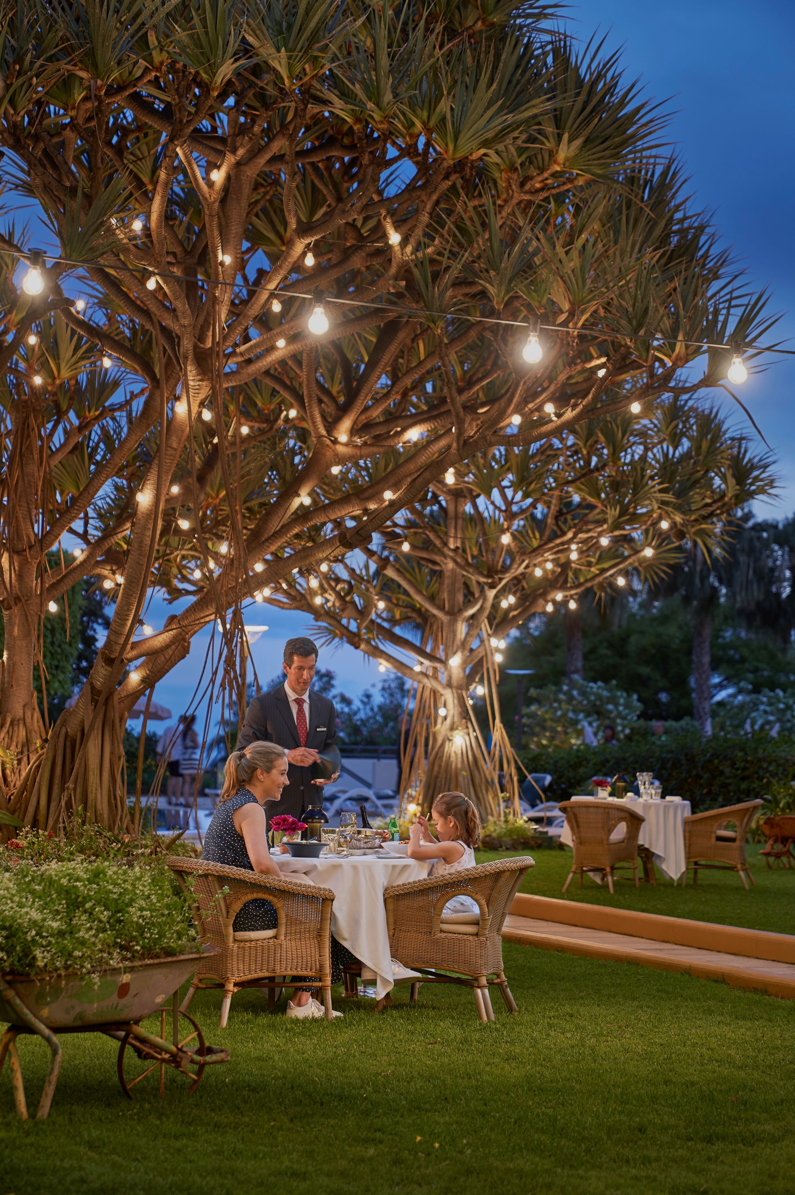 Un espace extérieur aménagé pour prendre les repas, avec une table dressée pour deux personnes, située sous un arbre décoré de guirlandes lumineuses.