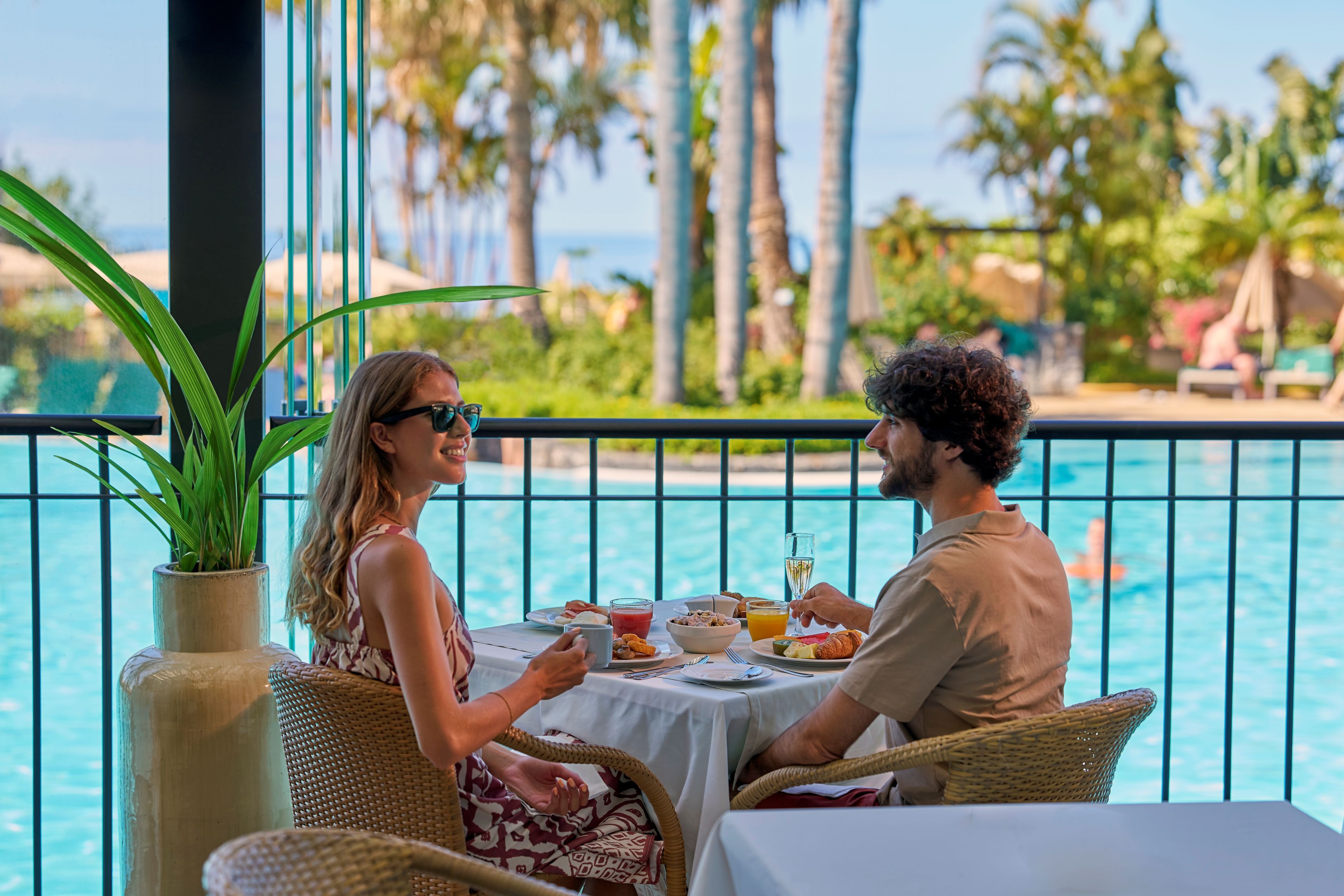A couple dining at a table with a view of a pool and palm trees.