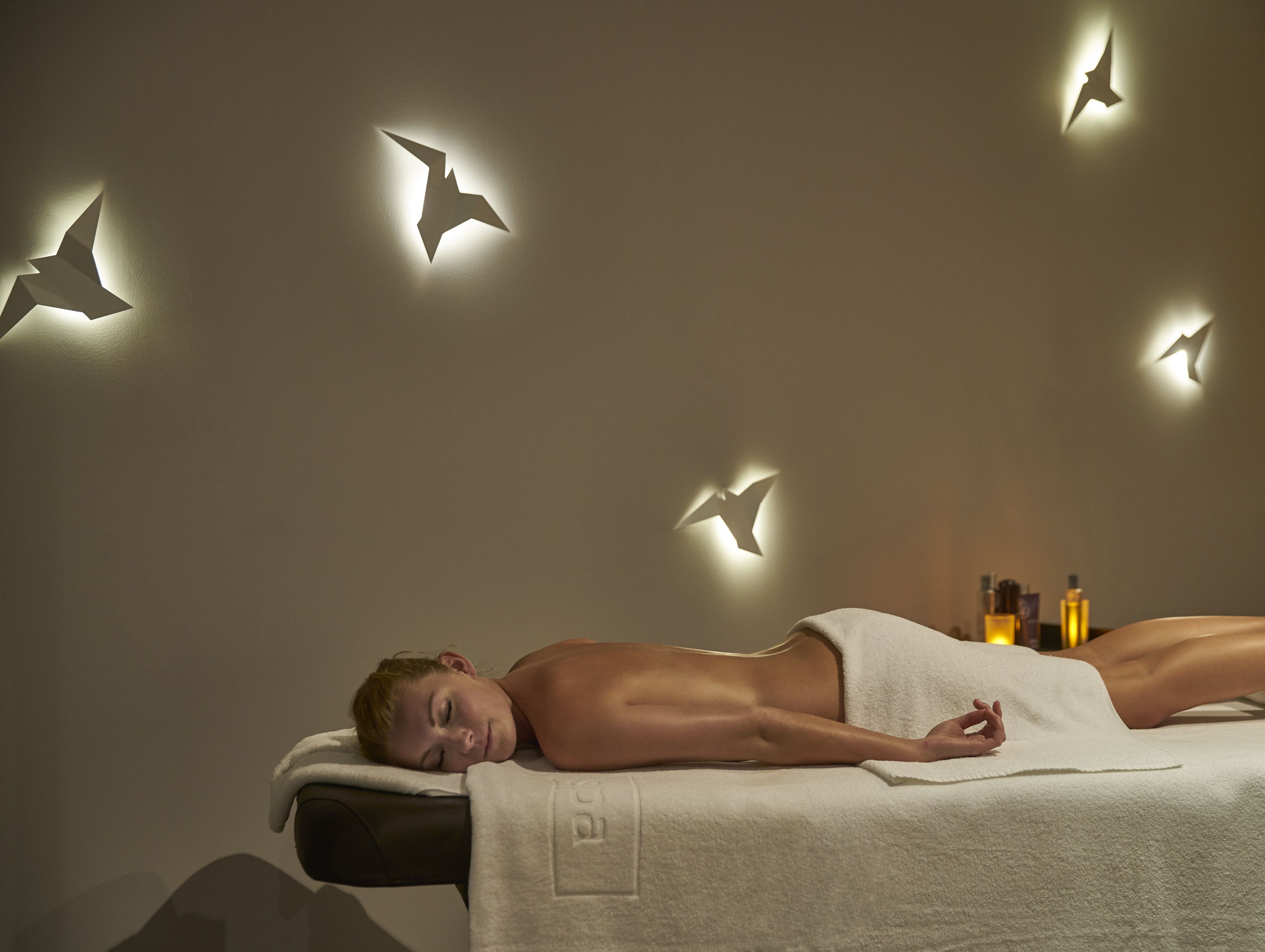 Um espaço de spa com uma pessoa deitada em uma mesa de tratamento, cercada por luminárias de parede com o formato de pássaros.