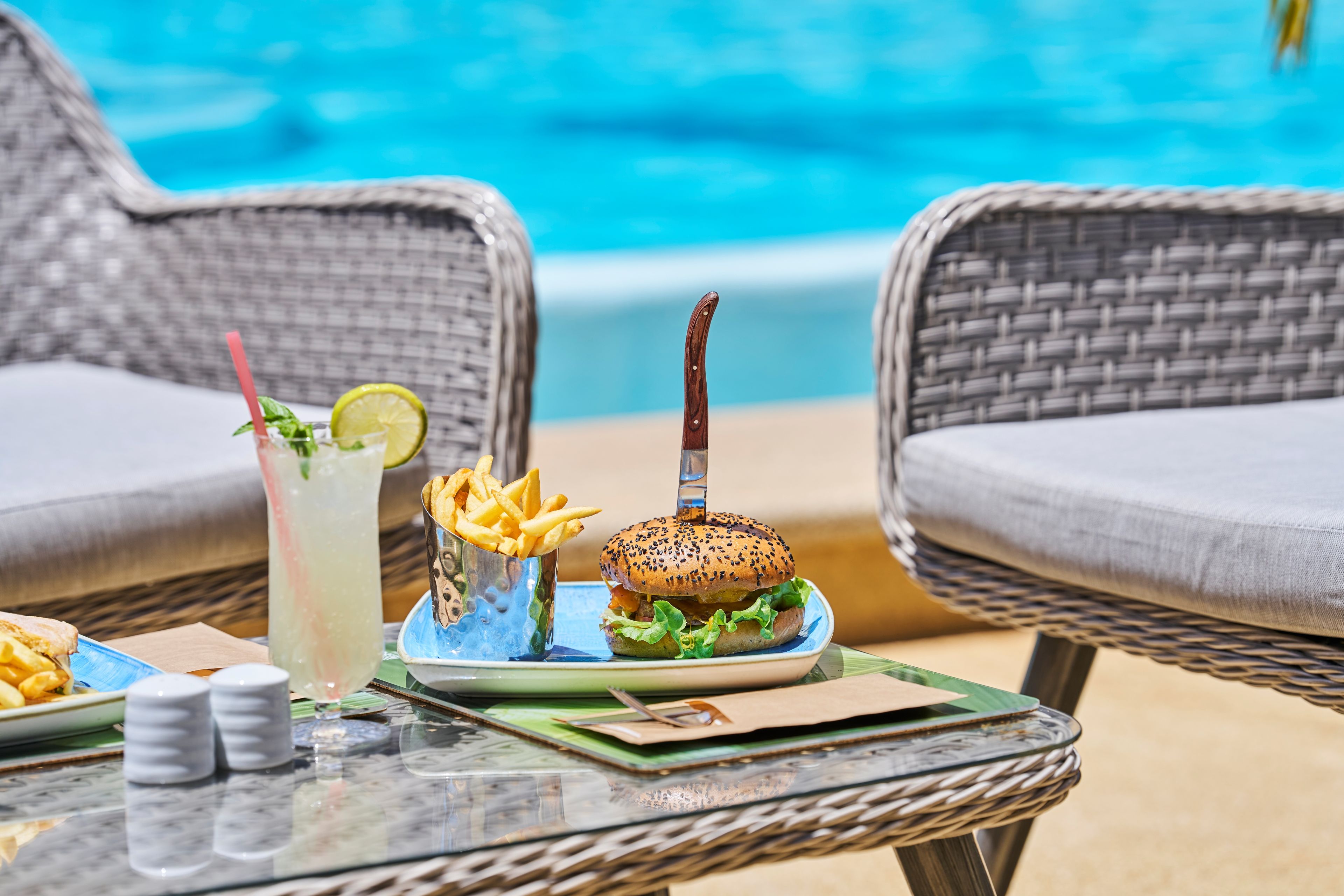 Une table à manger avec un hamburger, des frites et une boisson, le tout installé devant une piscine.