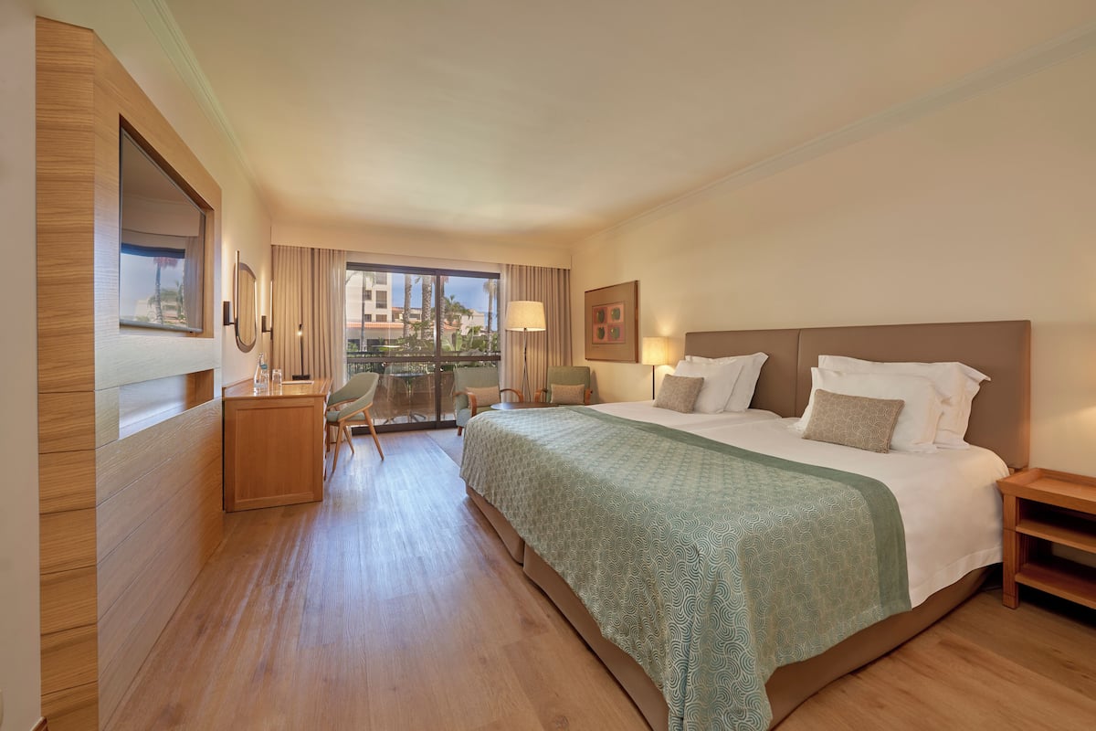 A hotel room with a large bed, a desk, a chair, and a balcony with a view of palm trees and buildings.
