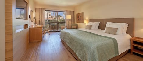 Habitación de hotel con una cama grande, un escritorio, una silla y un balcón con vistas a palmeras y edificios.