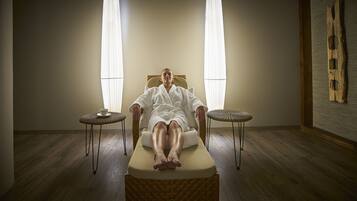A person lying on a massage table in a room with wooden flooring, two small round tables, and a large wooden wall decoration.