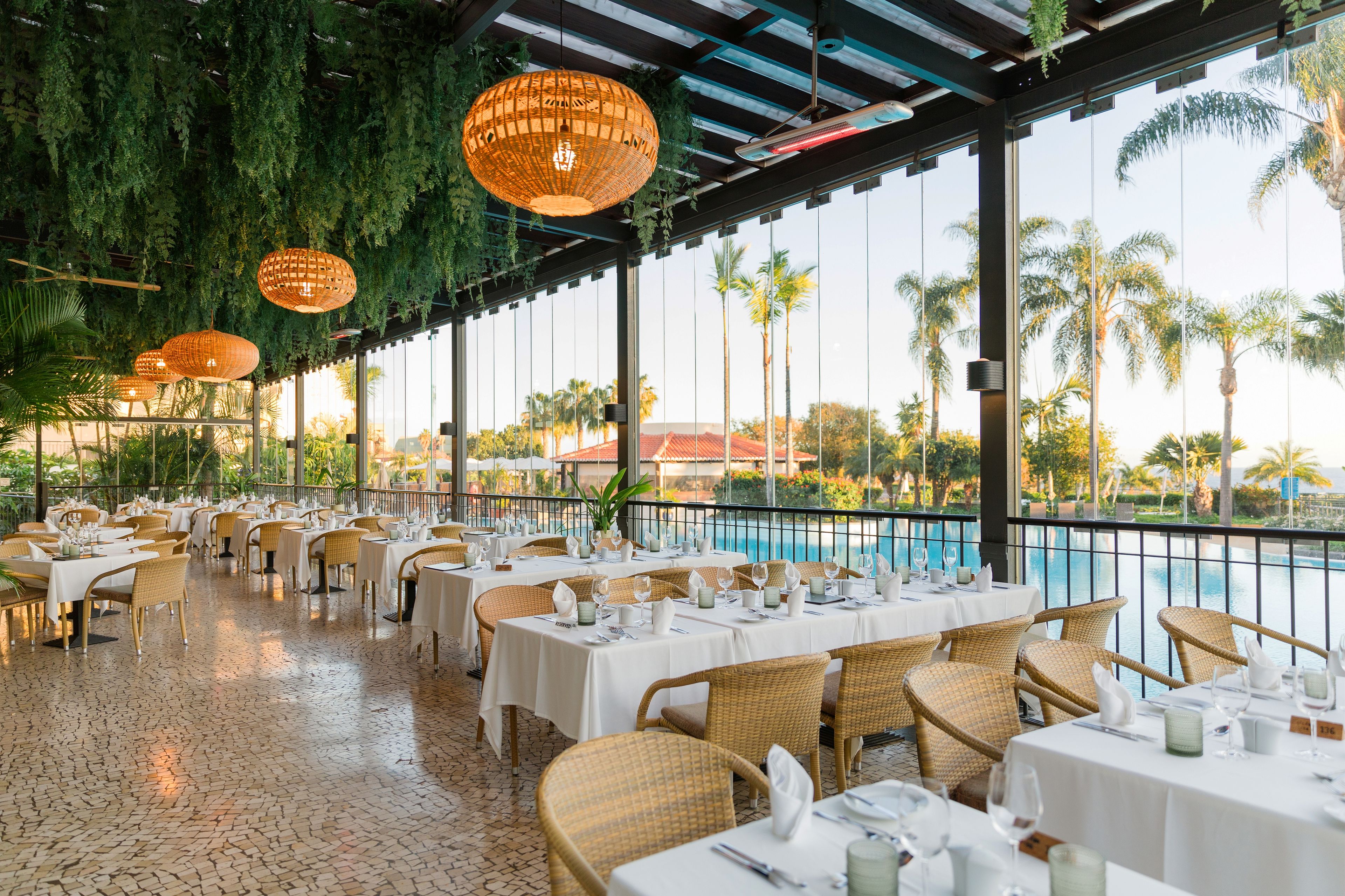 A restaurant with outdoor seating overlooking a pool, featuring wicker furniture and hanging lamps.