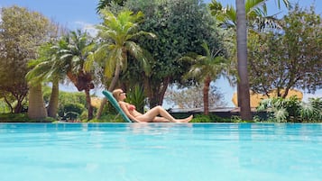 Eine Person entspannt auf einer Liege am Pool, umgeben von üppiger Vegetation und Palmen.