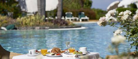 A dining table with breakfast items by a poolside, surrounded by lounge chairs and palm trees.