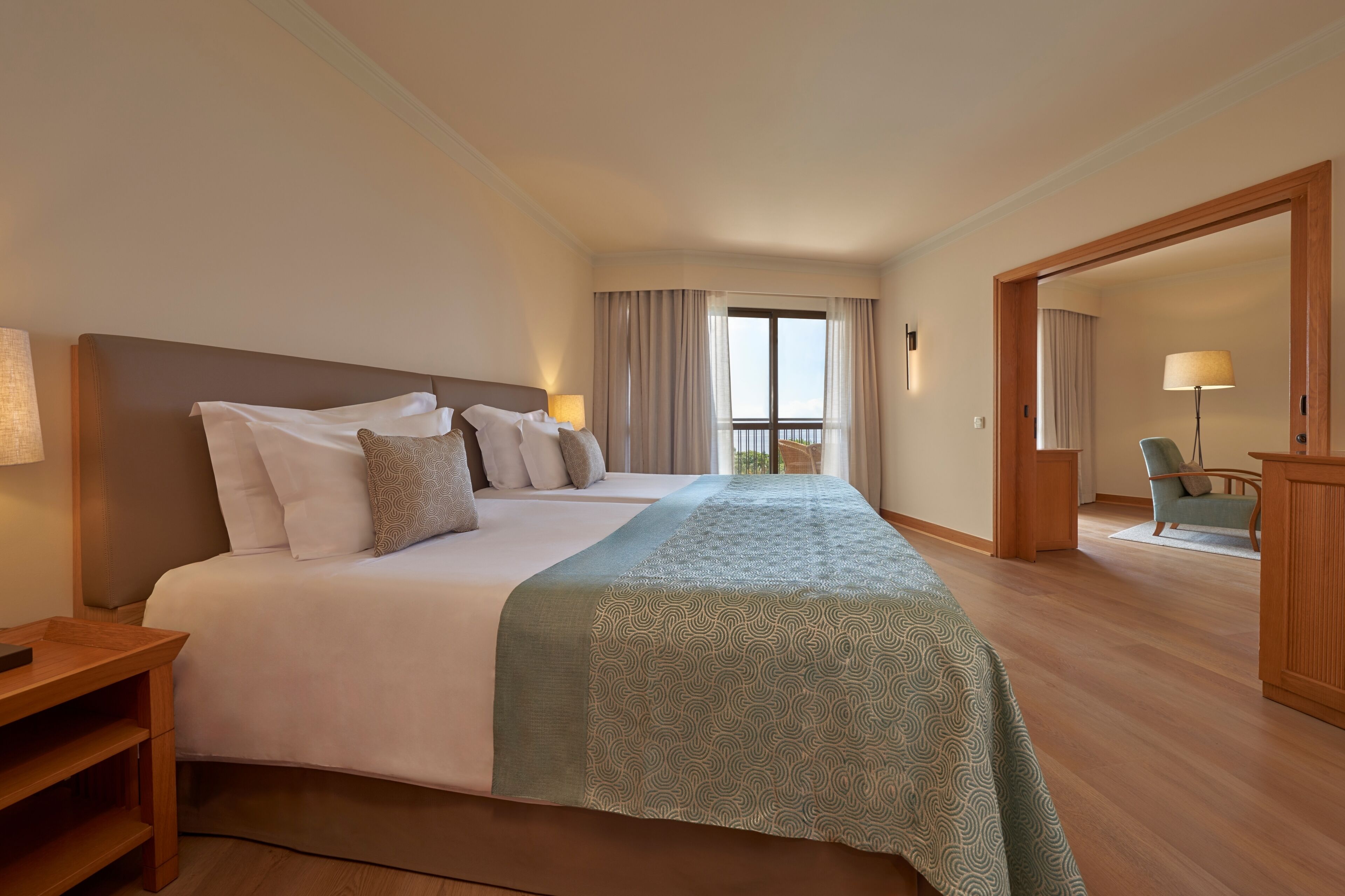 A hotel room with a large bed, a wooden nightstand, a chair, and a view of the outdoors through a window.