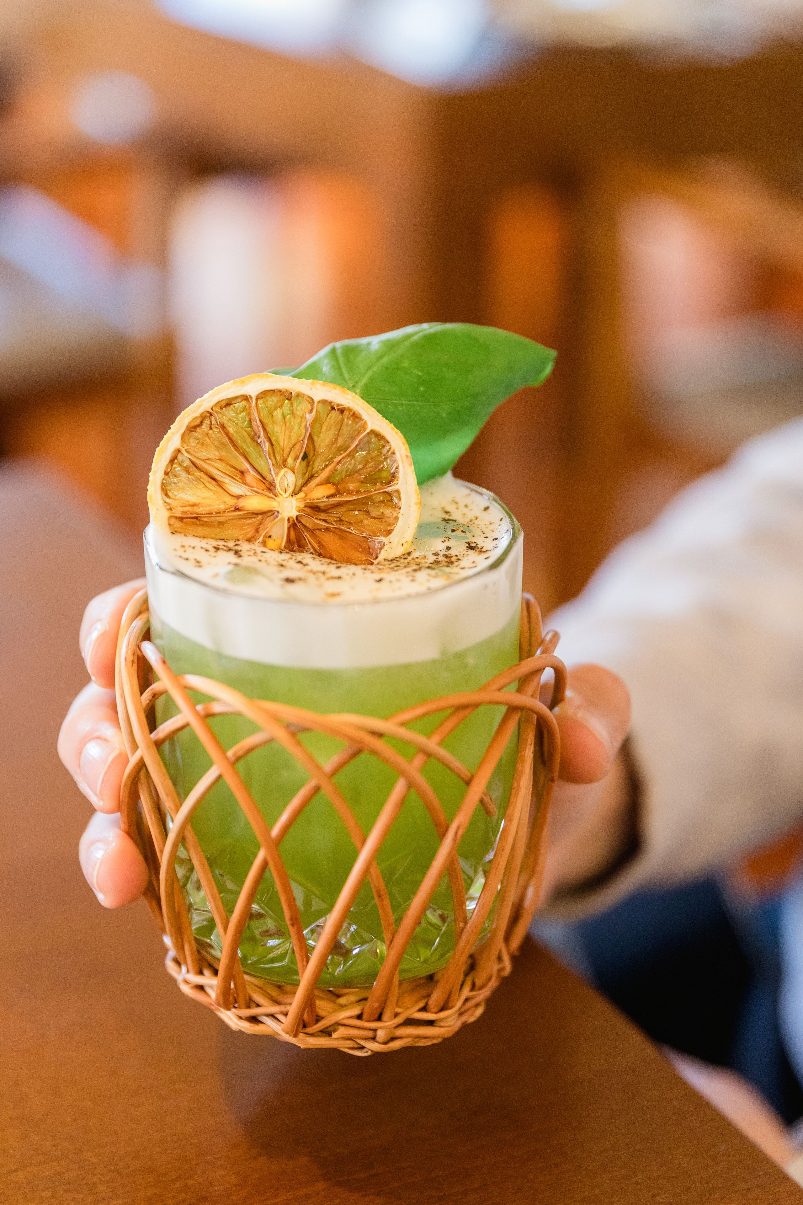 A person holding a glass with a layered drink, garnished with a dried orange slice and a leaf, in a woven basket.