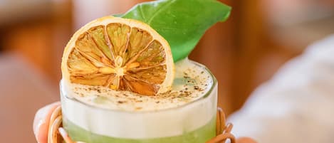 A person holding a glass with a layered drink, garnished with a dried orange slice and a leaf, in a woven basket.
