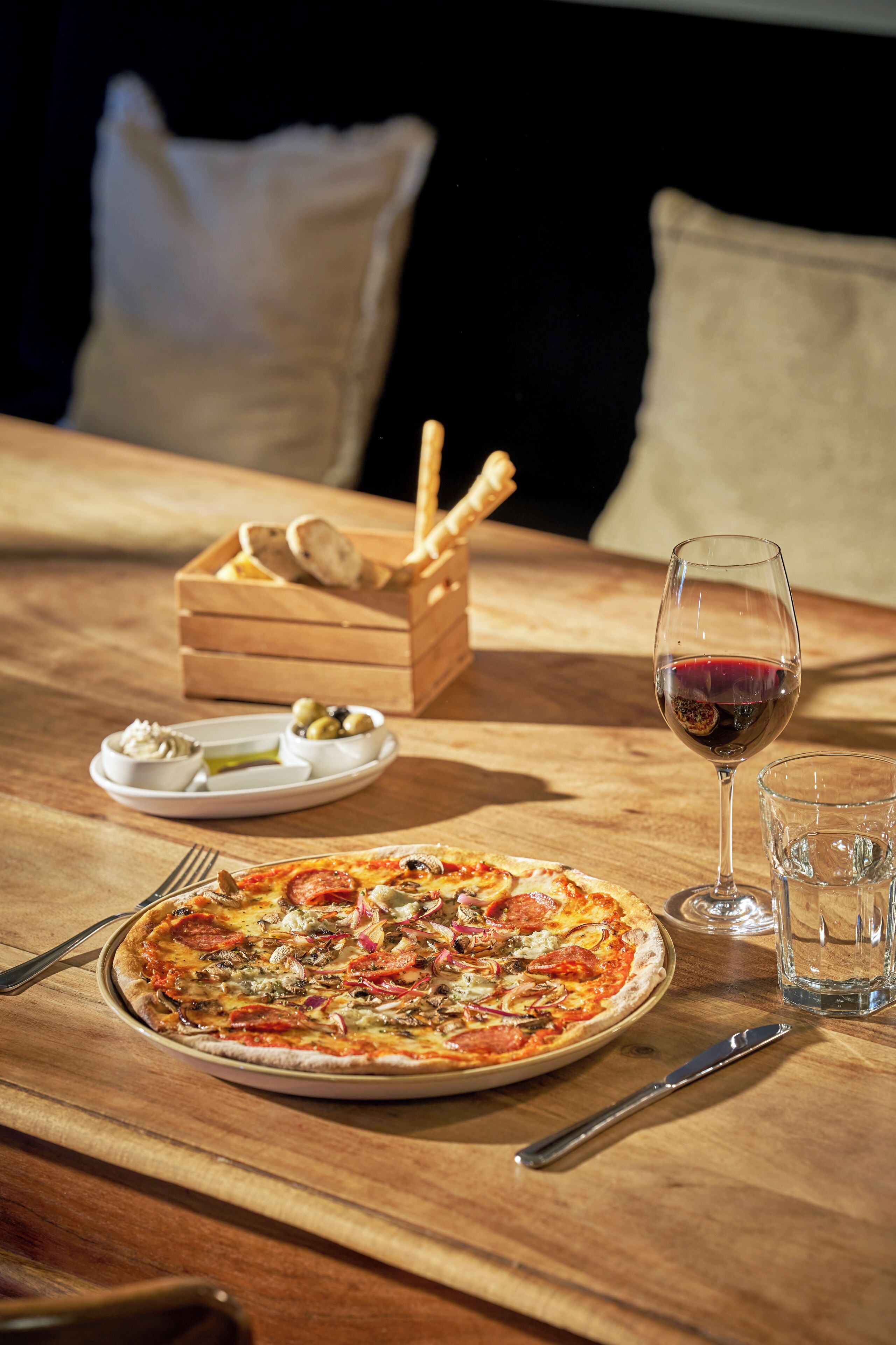A wooden table with a pizza, a glass of red wine, a bread basket, and a plate with olives and a small dish of sauce.