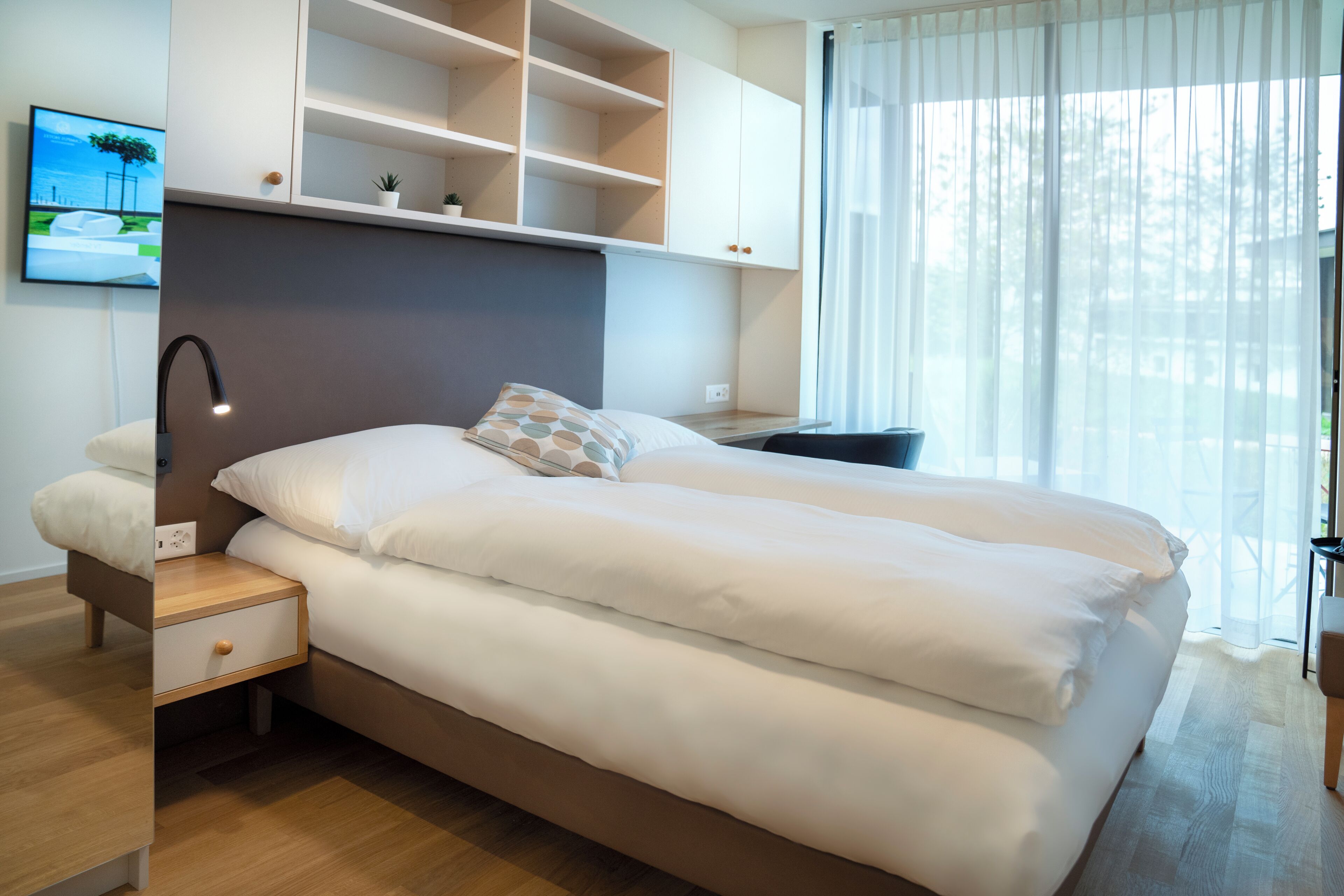 Um quarto moderno com cama, mesas de cabeceira, televisão e janela com cortinas leves.
