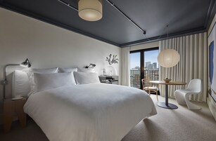 Ein modernes Hotelzimmer mit einem großen Bett, Nachttischen, einem Schreibtisch, einem Sessel und Blick auf die Stadt.