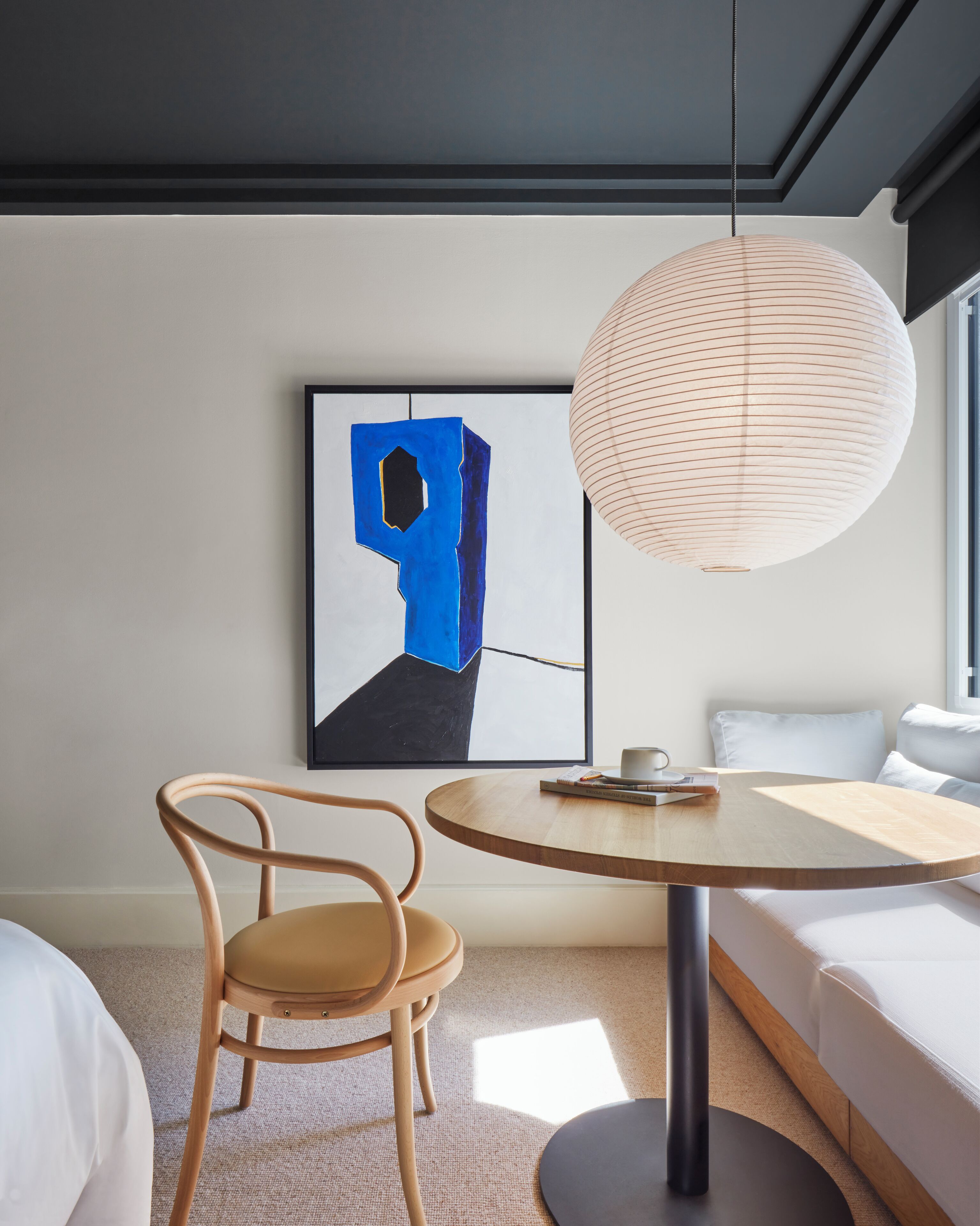 Ein modernes Zimmer mit einem runden Holztisch, einem blauen abstrakten Gemälde, einer gewobenen Leuchte, einem Holzstuhl und einem weißen Sofa.