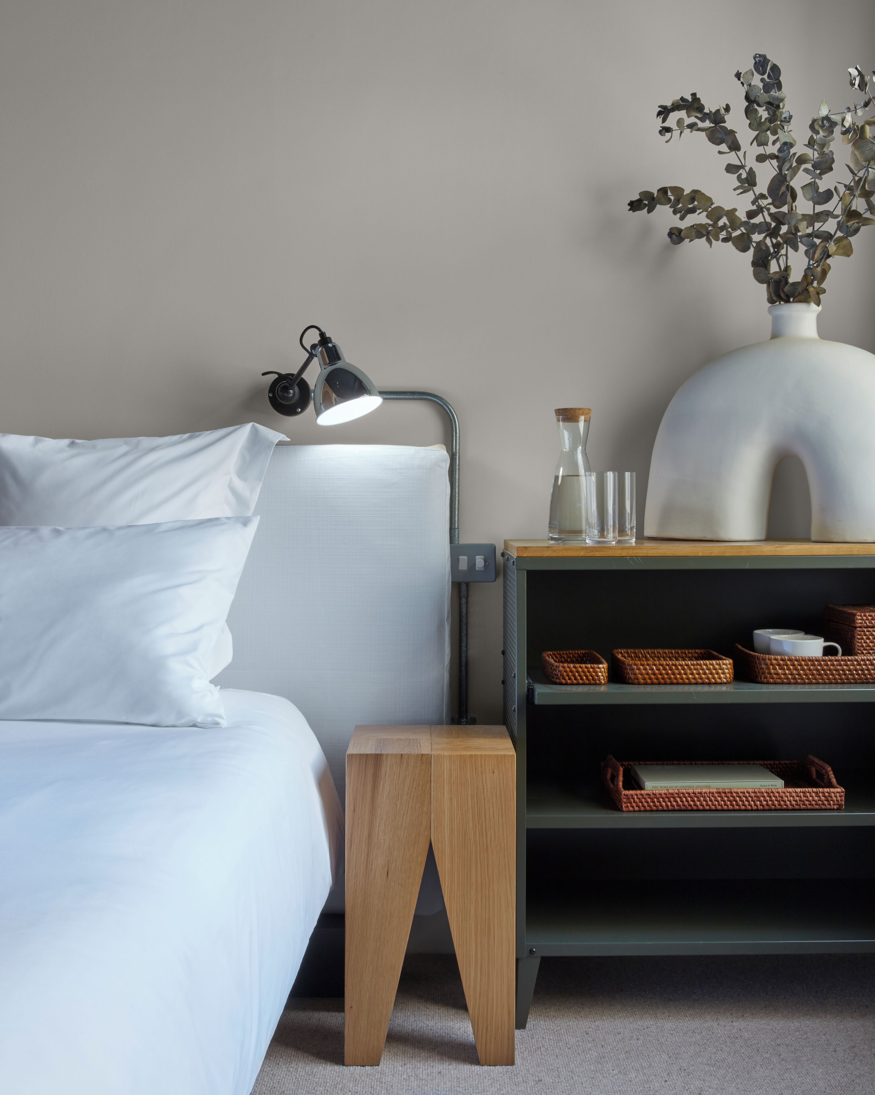 Un dormitorio ordenado con cama, lámpara de noche, mesita de noche con un florero y cestas de mimbre.