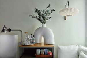 A modern living room with a white sofa, a wooden side table with a vase of green plants, a lamp, and a wall-mounted light fixture.