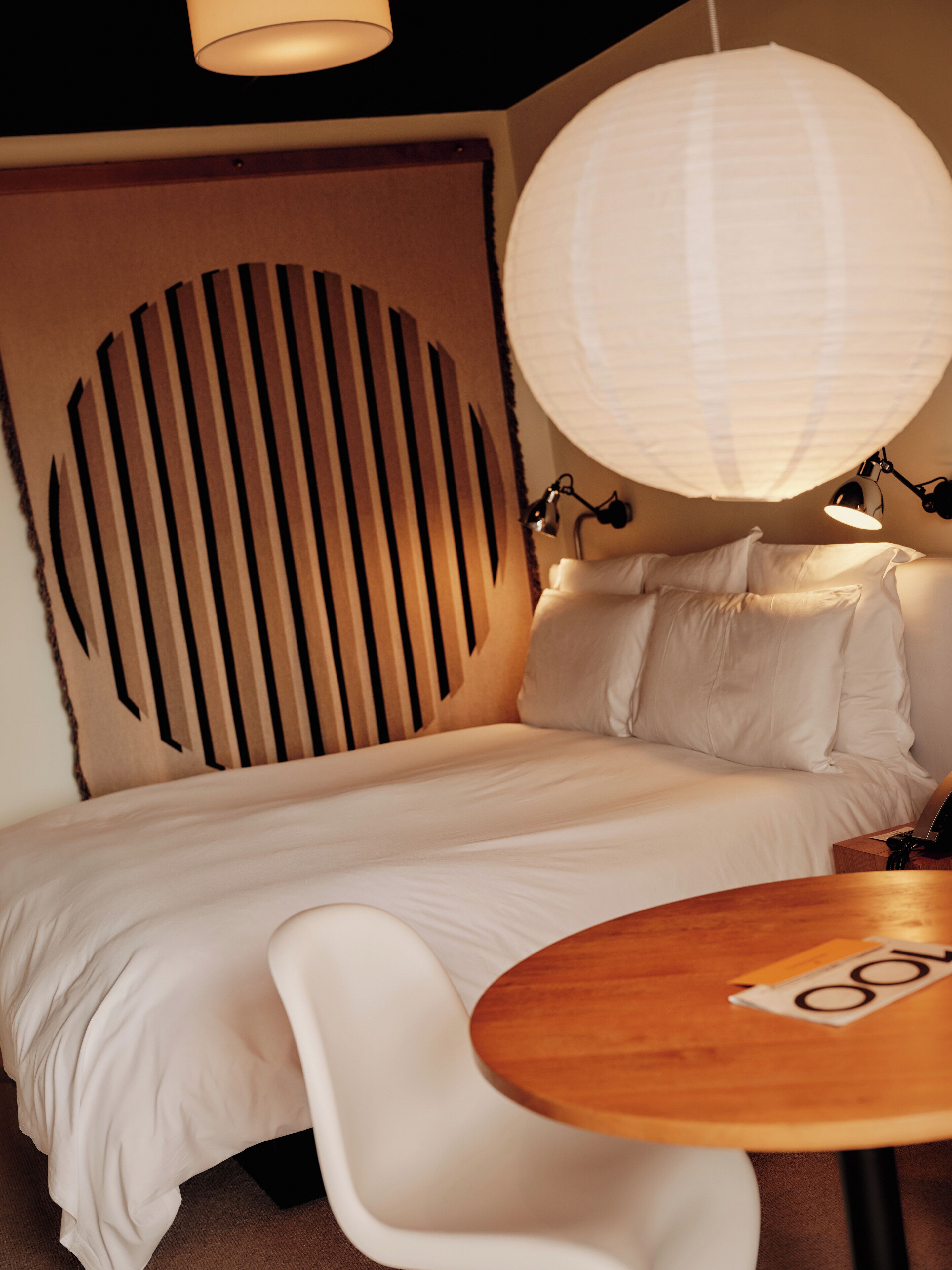 Un lit bien fait, avec du linge de lit blanc, une table ronde en bois surmontée d’un set de table, et une grande lanterne en papier blanc suspendue au-dessus.