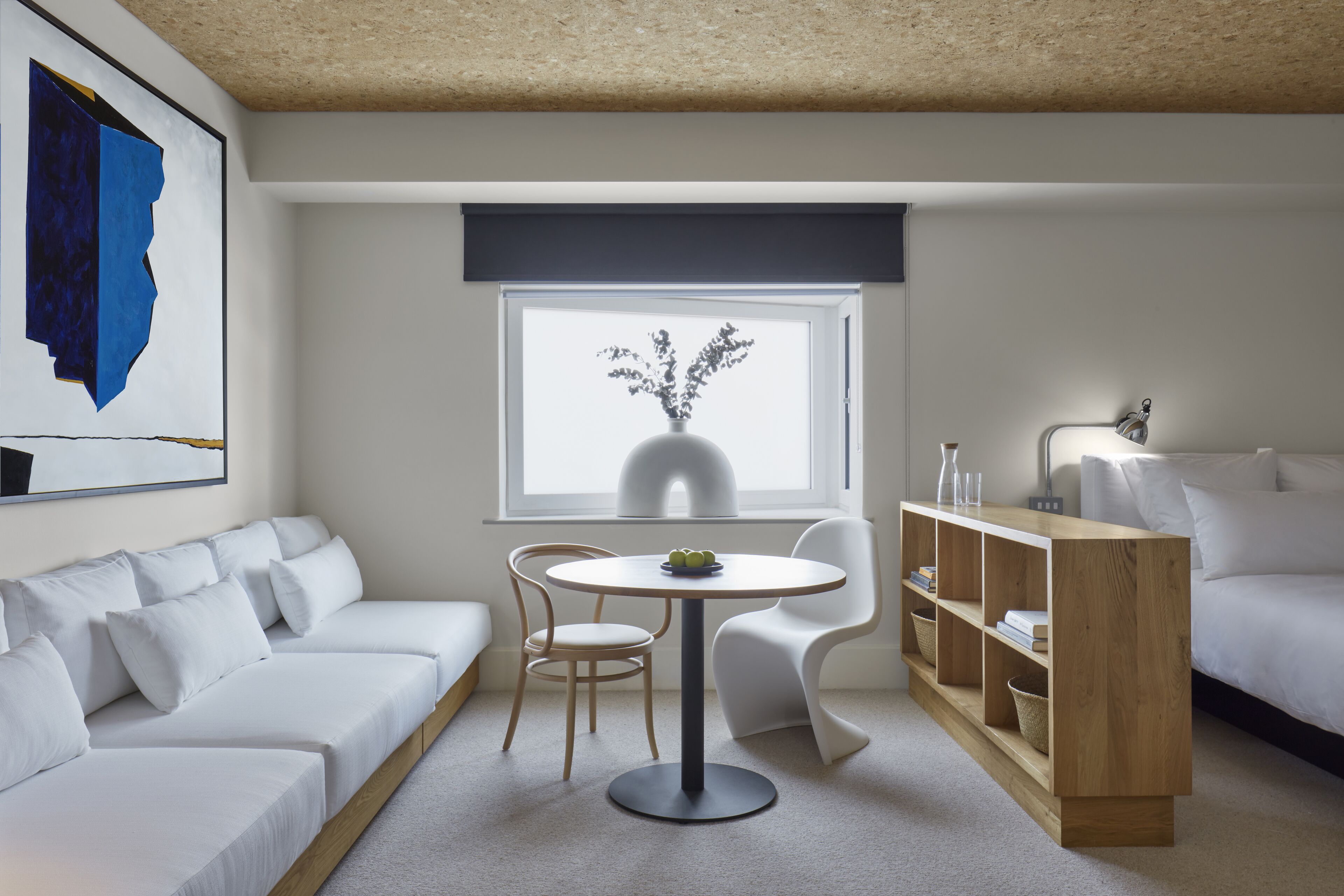 A modern room with a white sofa, a round table with a vase, a wooden bookshelf, and a large window.