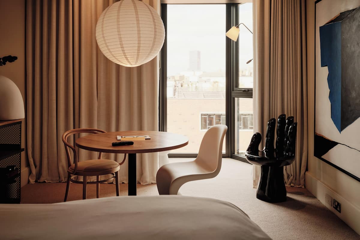 A hotel room with a bed, a round table with chairs, a large window with curtains, a hanging light fixture, and a black sculpture on the floor.