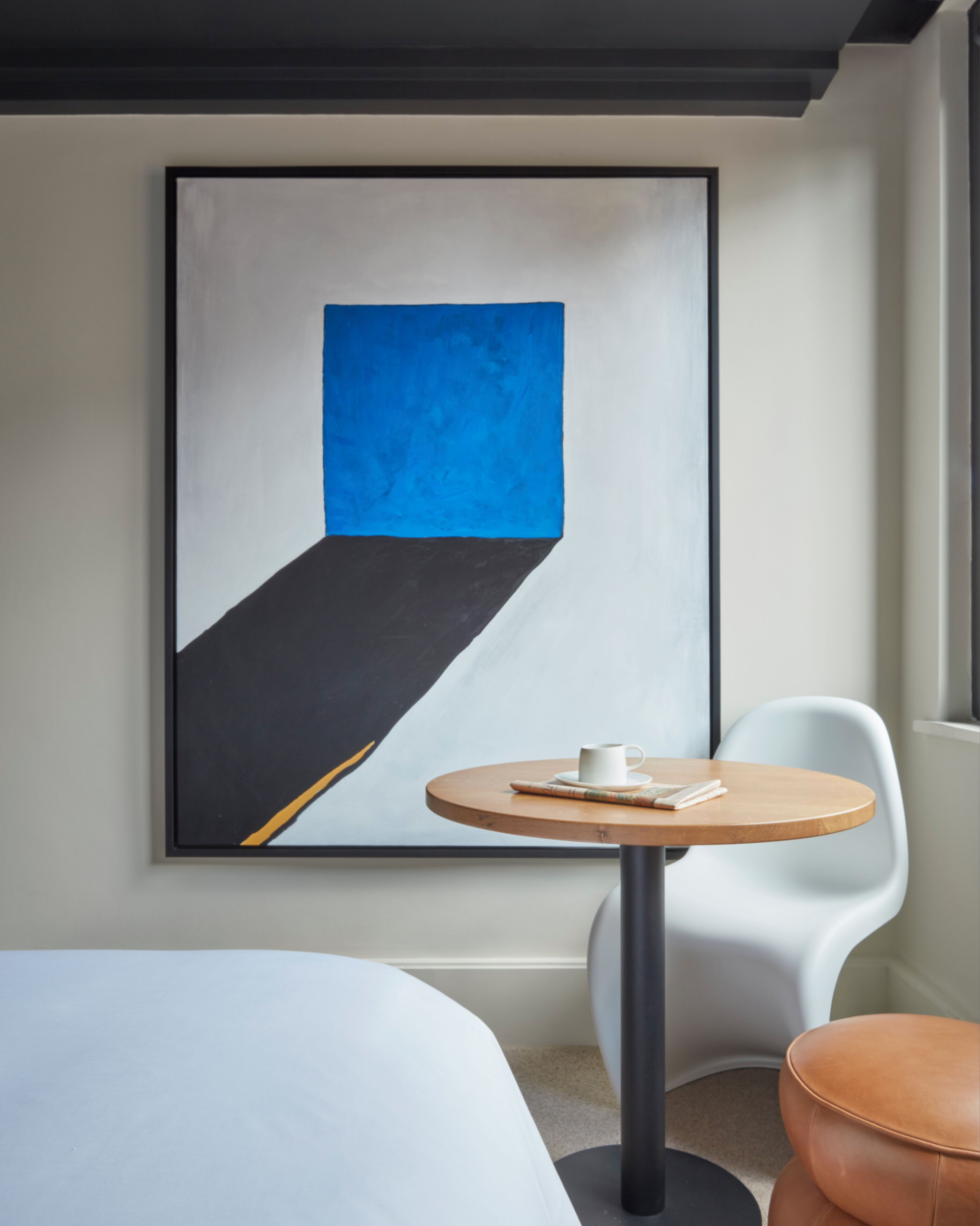 Ein Hotelzimmer mit einem großen abstrakten Gemälde an der Wand, einem runden Holztisch mit einer weißen Tasse, einem weißen Stuhl und einem braunen Lederhocker.