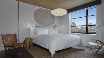 A modern bedroom with a large bed, two chairs, a bedside table, a hanging light fixture, and a view of the city through a large window.