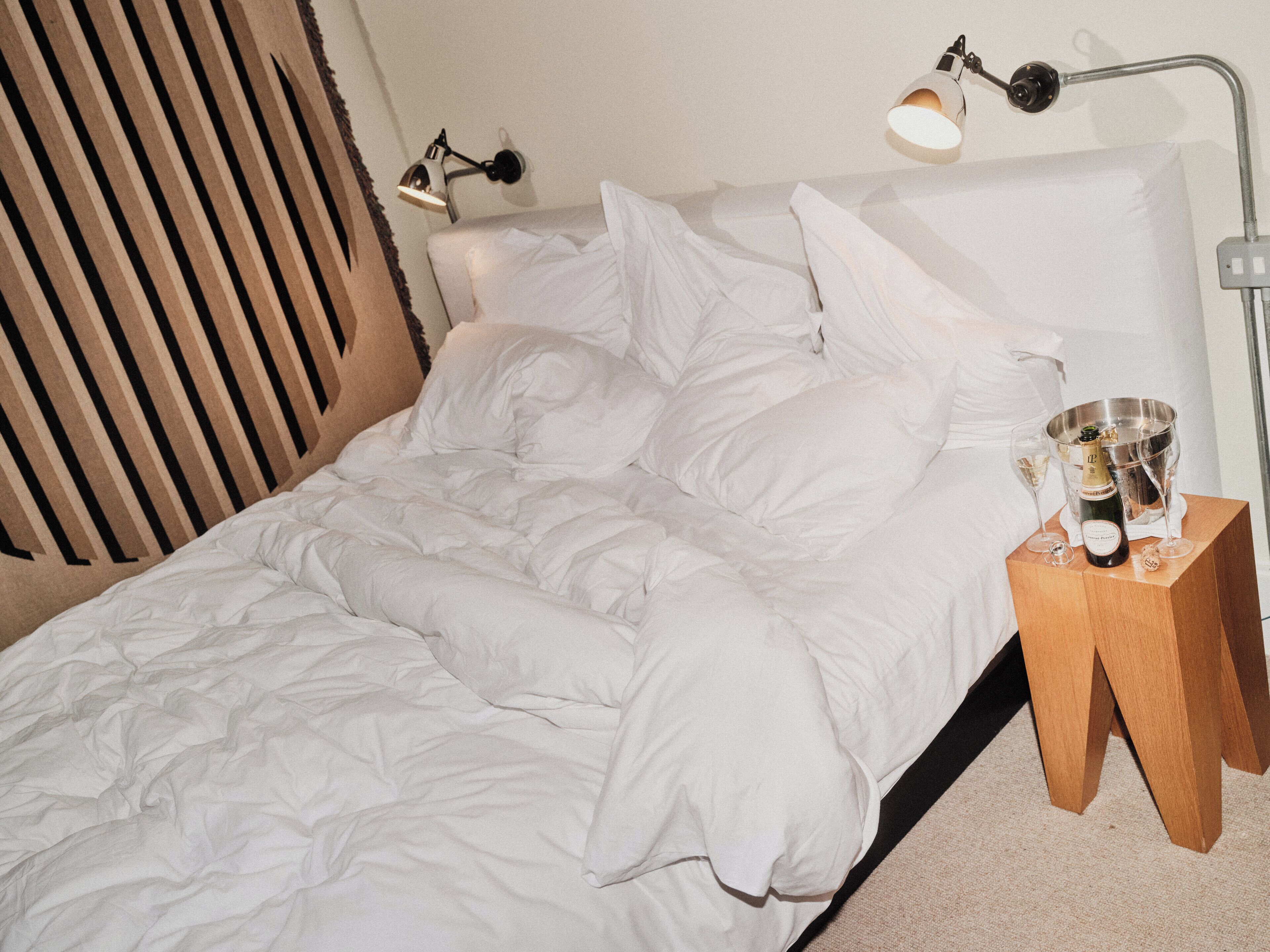 A neatly made bed with white linens, a bedside table with a bottle of champagne and glasses, and a wall-mounted lamp.