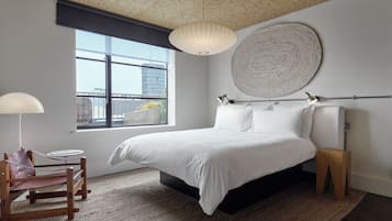 A modern bedroom with a large bed, a wooden chair, a round wall hanging, and a view of the city through a window.