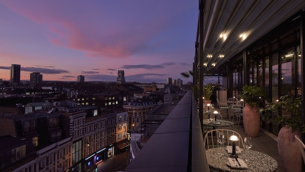 A balcony with a city view at dusk, featuring tables with chairs and potted plants. - One Hundred Shoreditch (London)