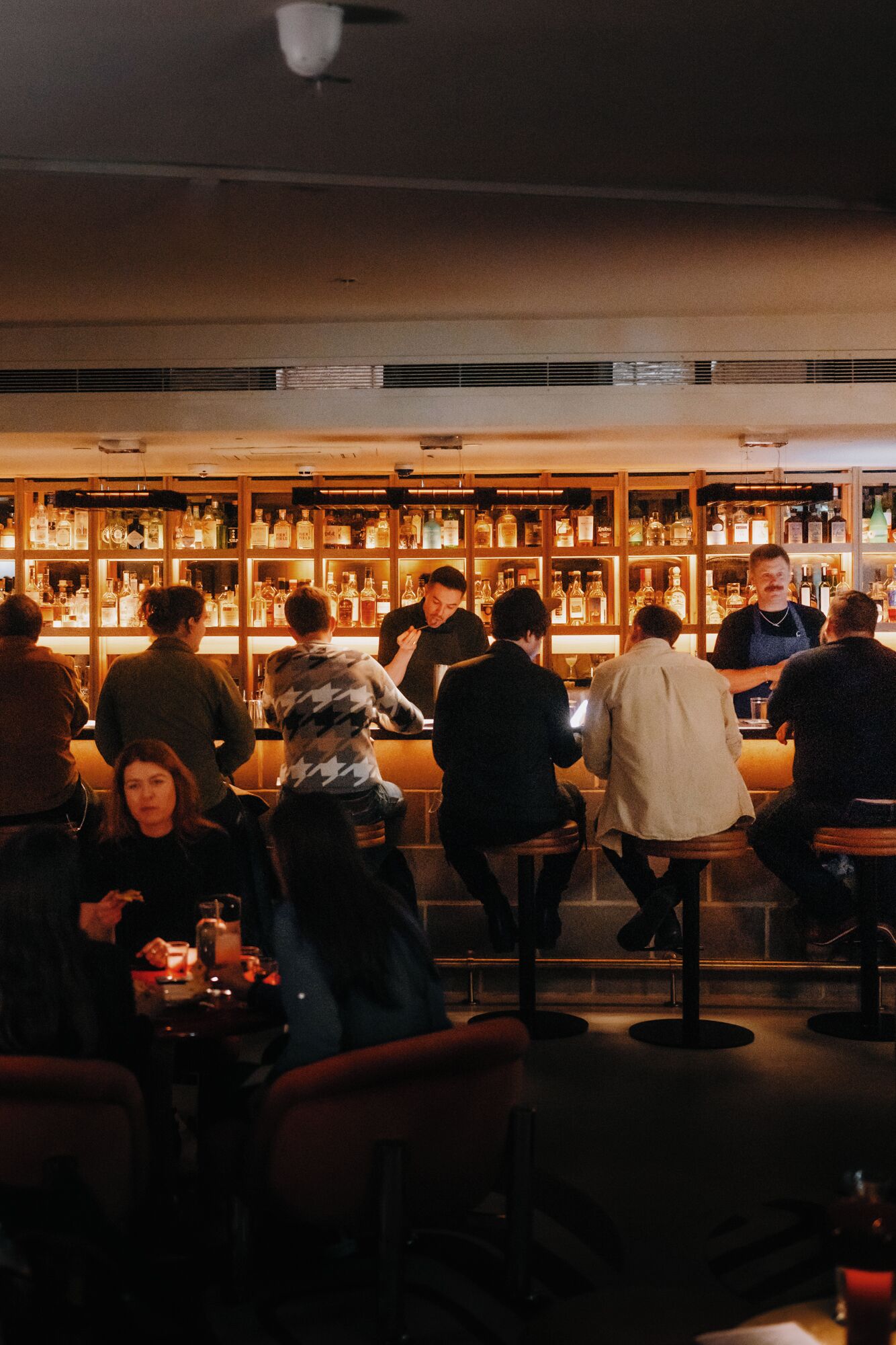 Eine Bar mit Gästen an Tischen, einem Barkeeper, der Getränke ausschenkt, und Regalen voller Flaschen.