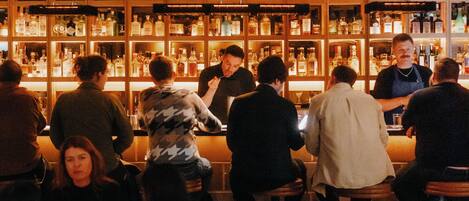 Eine Bar mit Gästen an Tischen, einem Barkeeper, der Getränke ausschenkt, und Regalen voller Flaschen.