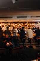 A bar with patrons seated at tables, a bartender serving drinks, and shelves stocked with bottles behind the bar.