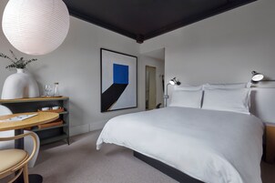 Una habitación de hotel moderna con una cama grande, un escritorio con una silla, un jarrón con plantas, un cuadro abstracto enmarcado y una lámpara colgante.