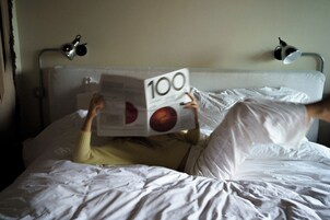 Eine Person liegt im Bett, ein Buch mit dem Titel '100' im Gesicht.