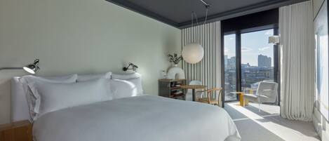 A modern hotel room with a large bed, a desk, a chair, and a view of the city.