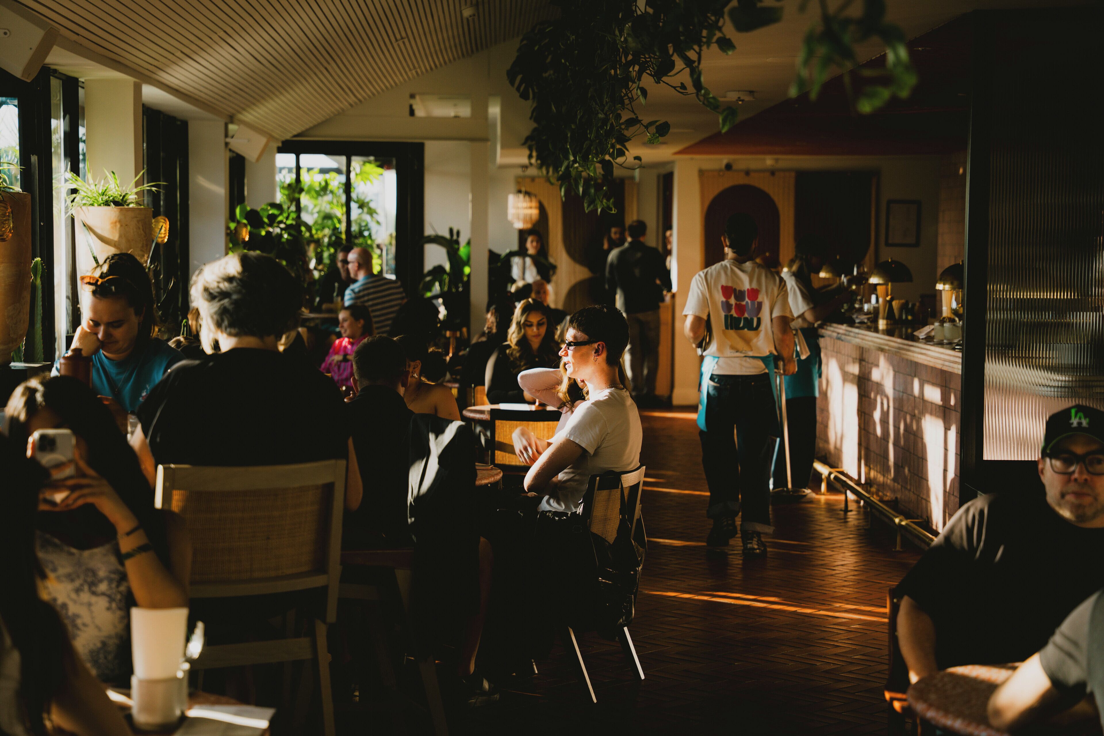 A busy indoor dining area with wooden flooring, potted plants, and a bar counter.