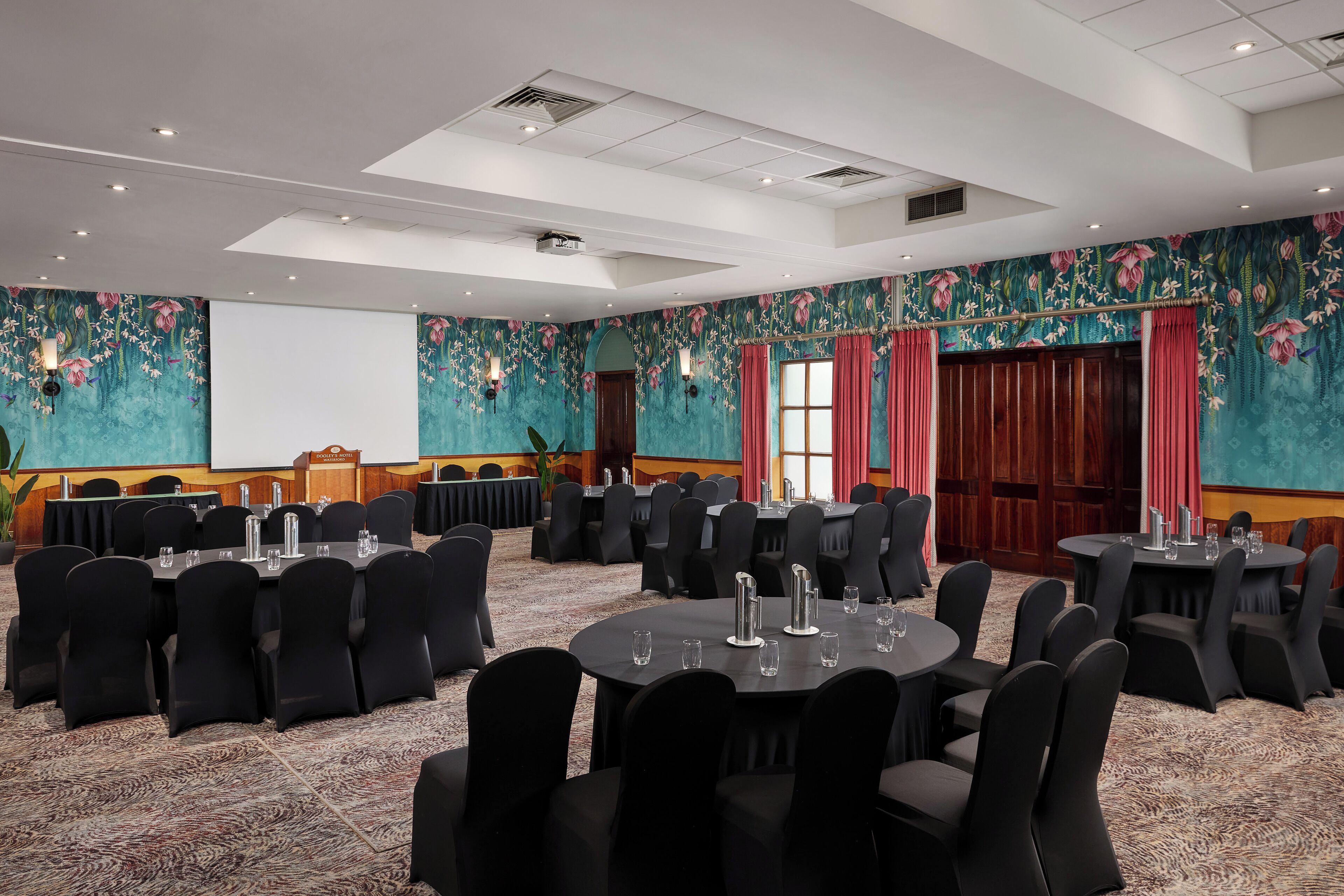 A conference room with round tables and black chairs, a stage with a podium, and floral wall decorations.