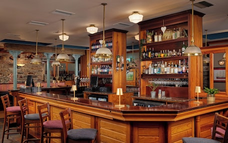 Zona de bar con mostradores de madera, diversas opciones de asiento y una variedad de botellas y vasos en estantes.