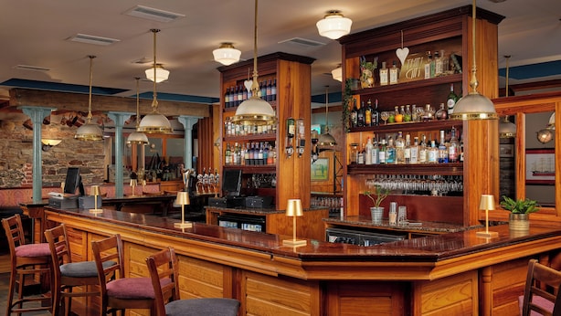 Zona de bar con mostradores de madera, diversas opciones de asiento y una variedad de botellas y vasos en estantes.