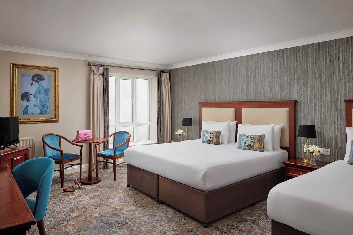 A hotel room with a large bed, two bedside tables, a desk with a chair, a small table with a chair, and a window with curtains.