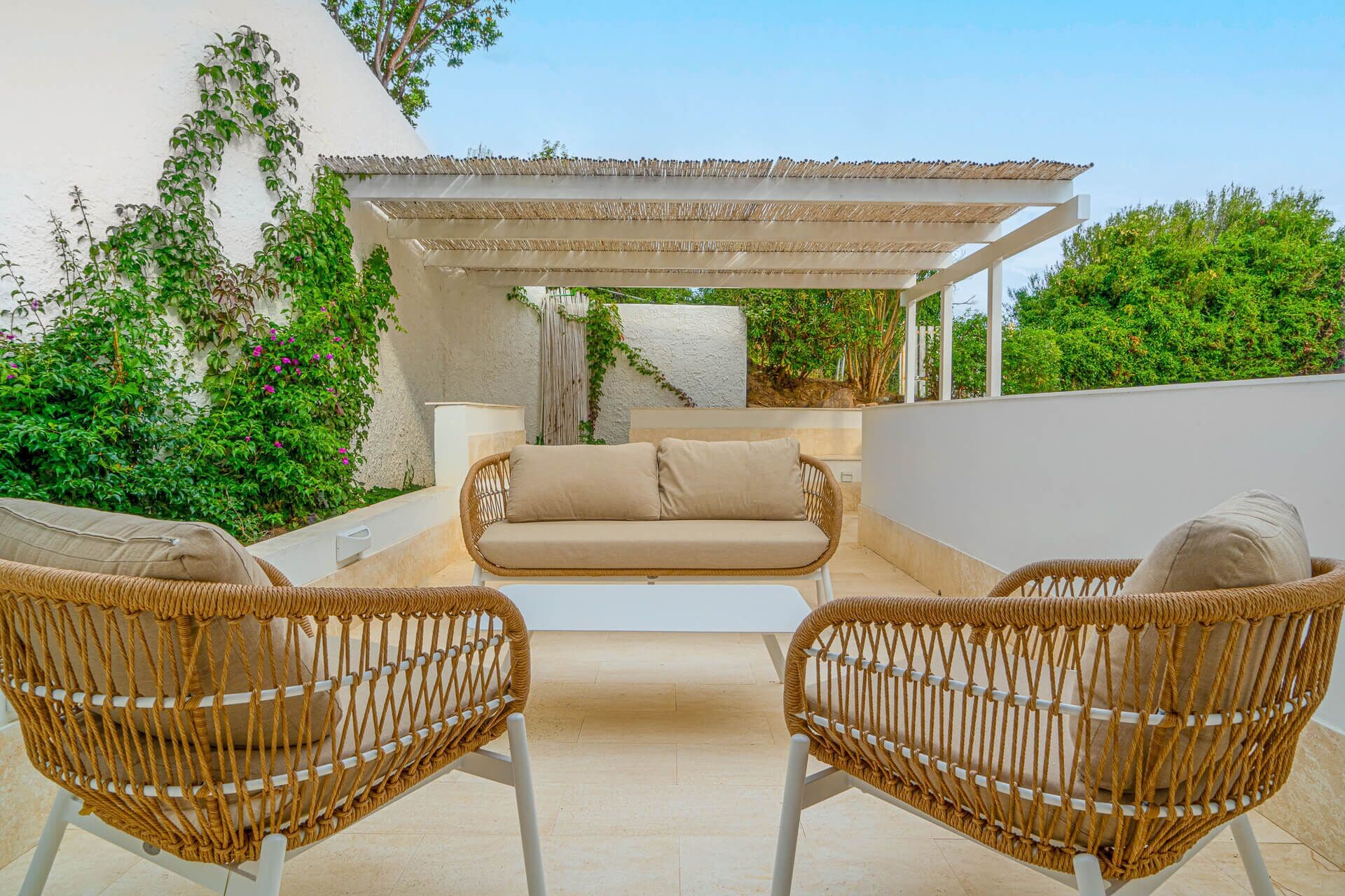 Ein Sitzbereich im Freien mit Korbstühlen, einer Couch, einer Pergola und üppigem Grün.