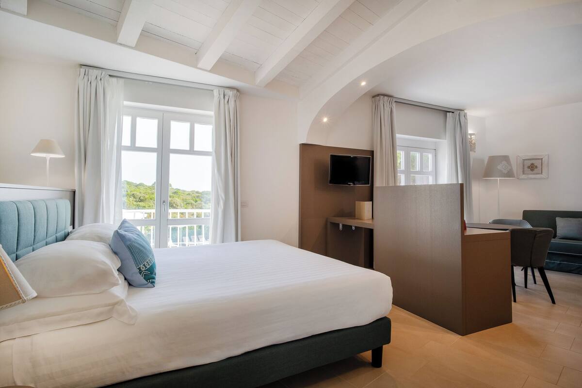 Ein modernes Hotelzimmer mit einem großen Bett, einem Schreibtisch, einem Fernseher und einem Balkon mit Vorhängen.