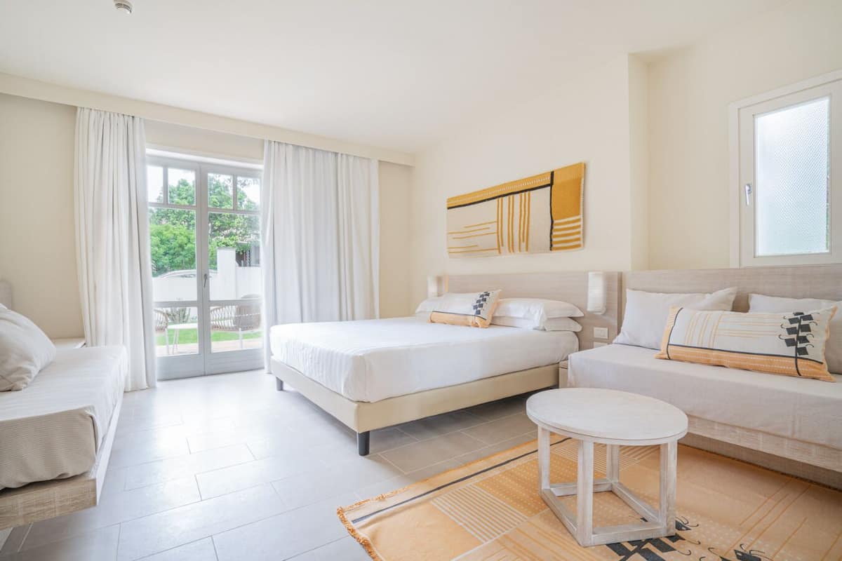 Ein modernes Hotelzimmer mit einem großen Bett, einer Couch, einem kleinen Tisch und Blick ins Freie durch eine Schiebetür.
