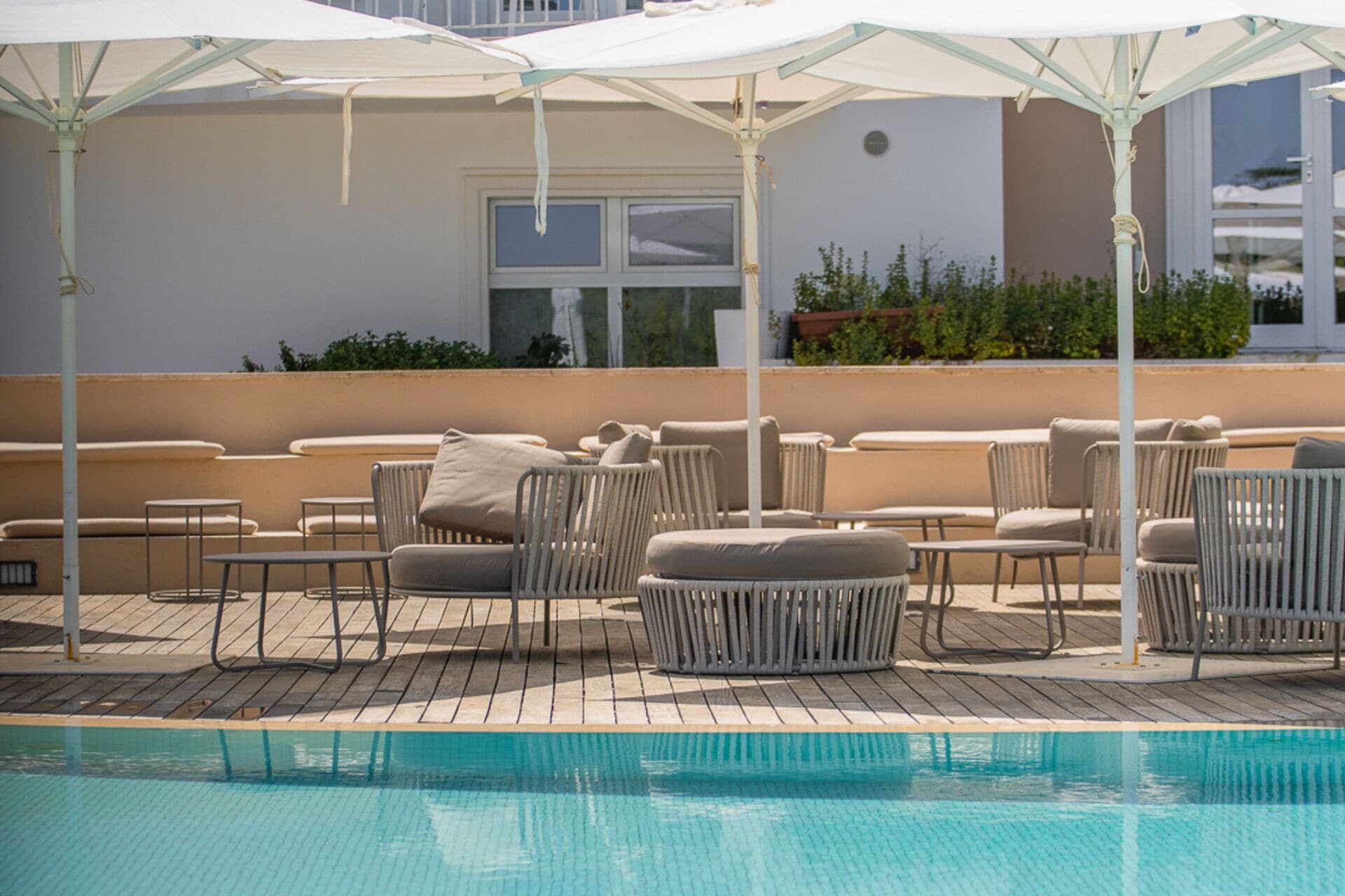 A rooftop pool area with lounge chairs and umbrellas, a pool, and a building with a window and greenery in the background.
