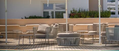 A rooftop pool area with lounge chairs and umbrellas, a pool, and a building with a window and greenery in the background.
