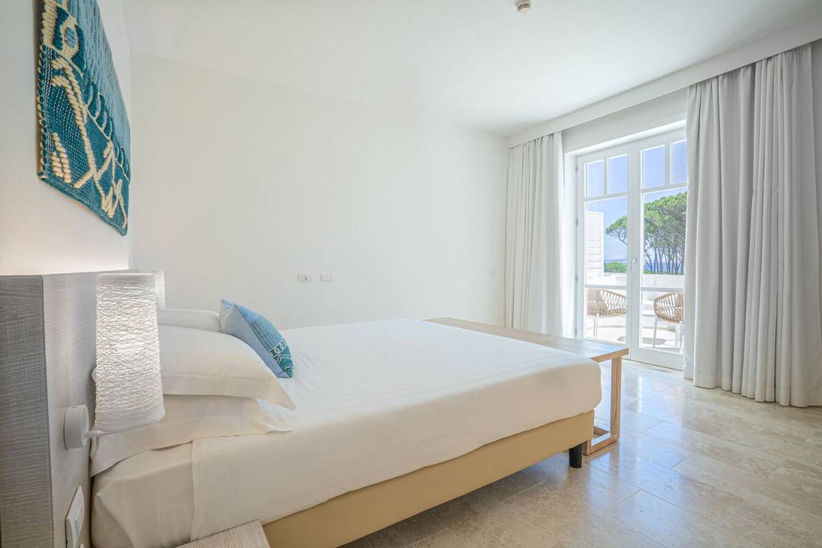 Ein Hotelzimmer mit Bett, Nachttisch, Lampe und Blick ins Freie.
