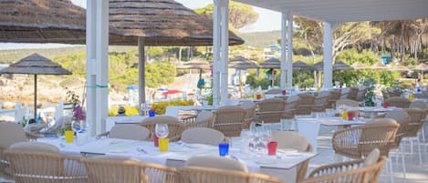 Un restaurant en plein air avec des tables blanches et des chaises en osier, entouré de parasols en chaume et offrant une vue magnifique sur la plage.