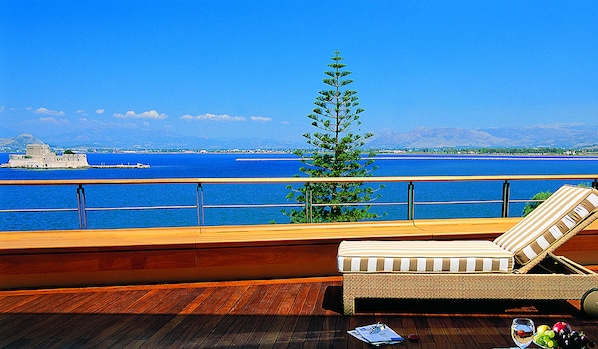 A balcony with a wooden floor, a lounge chair, and a view of the sea and a distant island.