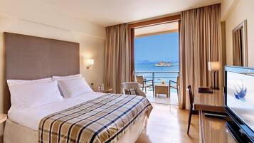 A hotel room with a bed, a desk, a TV, and a balcony with a view of the sea.