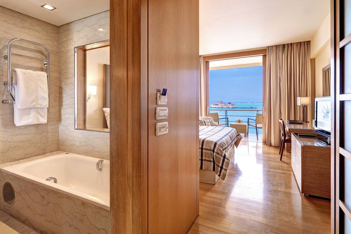Una habitación de hotel moderna con una bañera grande, un balcón con vista al mar, un escritorio con computadora y una cama con colcha a cuadros.